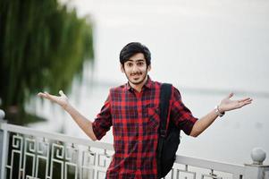 jeune étudiant indien en chemise à carreaux et jeans avec sac à dos posé sur la ville du soir contre le lac, montre l'émotion. photo