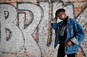 homme afro-américain en veste de jeans, béret et lunettes contre le mur de briques de graffiti avec le signe bruk. photo