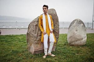 homme élégant indien en vêtements traditionnels jaunes avec foulard blanc posé en plein air contre de grosses pierres. photo