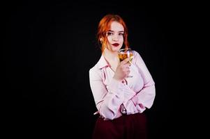 magnifique fille aux cheveux rouges en chemisier rose et jupe rouge avec un verre de vin à portée de main isolé sur noir. photo