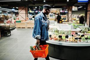 homme afro-américain décontracté et élégant à la veste en jean et au béret noir tenant un panier avec des ananas dans la section bio des fruits du supermarché. photo