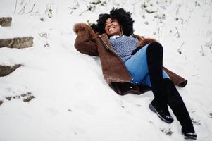 cheveux bouclés femme afro-américaine porter sur un manteau en peau de mouton et des gants posés le jour de l'hiver, se trouve dans la neige. photo