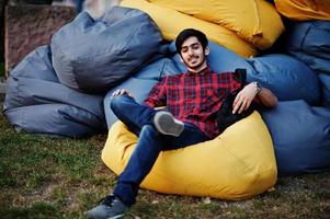 jeune étudiant indien en chemise à carreaux et jeans assis et se détendre sur des oreillers extérieurs. photo