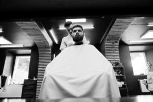 jeune homme barbu se coupe les cheveux par un coiffeur assis sur une chaise au salon de coiffure. âme de barbier. photo