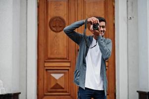 Un jeune photographe indien élégant porte une tenue décontractée posée en plein air avec un appareil photo reflex numérique à portée de main.