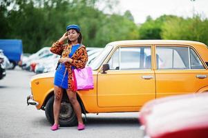 belle dame afro-américaine avec des sacs à provisions debout près d'une voiture rétro classique orange. photo