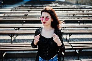 portrait de jeune fille brune à lunettes roses porter sur noir posé en plein air par une journée ensoleillée contre une rangée de bancs. photo