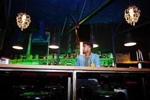 portrait d'un beau sud-asiatique barbu réussi, jeune pigiste indien en chemise de jeans bleu assis dans une boîte de nuit contre un comptoir de bar avec un cocktail et se reposer. photo