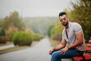 grand homme à la barbe arabe à la mode portant une chemise, un jean et des lunettes de soleil assis sur un banc rouge au parc. photo