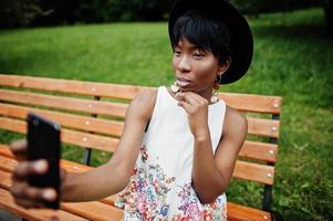 incroyable femme modèle afro-américaine en pantalon vert et chapeau noir posé sur un banc au parc et faisant selfie sur téléphone mobile. photo