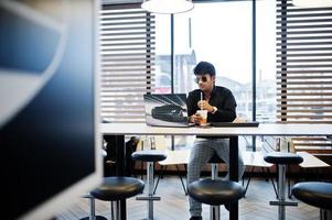 homme indien élégant à lunettes de soleil assis au café de restauration rapide contre son ordinateur portable. photo