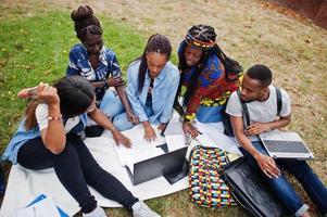 groupe de cinq étudiants africains qui passent du temps ensemble sur le campus de la cour universitaire. amis afro noirs assis sur l'herbe et étudiant avec des ordinateurs portables. photo