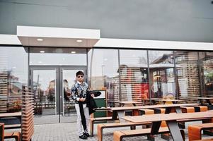 séduisant et beau homme indien élégant portant une chemise et des lunettes de soleil debout sur la terrasse du café-salon moderne lors de sa pause de travail. photo