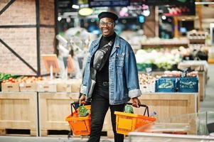 élégant homme afro-américain décontracté à la veste en jean et au béret noir tenant deux paniers, marchant et faisant du shopping au supermarché. photo