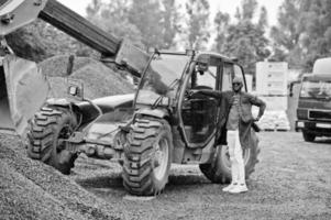 élégant homme afro-américain en chapeau et lunettes de soleil posé en plein air sous la pluie contre un tracteur avec un seau. photo