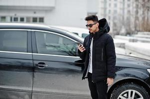 jeune homme indien décontracté en veste noire et lunettes de soleil posées contre une voiture suv avec un téléphone à portée de main. photo