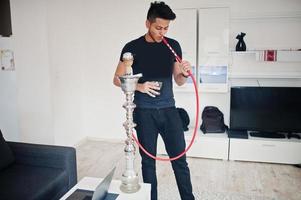 homme indien beau et à la mode en noir debout dans la chambre, fumant le narguilé avec des lunettes à portée de main. photo