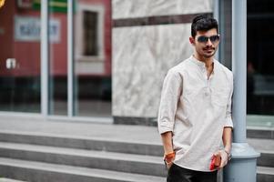 homme modèle indien élégant dans des vêtements décontractés et des lunettes de soleil posés en plein air dans la rue de l'inde. photo