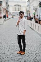 homme modèle indien élégant dans des vêtements décontractés et des lunettes de soleil posés à l'extérieur dans la rue de l'inde et parlant au téléphone mobile. photo