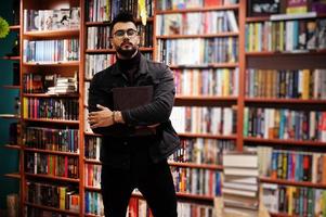 grand étudiant arabe intelligent, portant une veste en jean noir et des lunettes, à la bibliothèque avec un livre à portée de main. photo