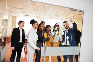 équipe de jeunes multiculturels pointant sur du verre avec des notes de papier colorées. groupe diversifié d'employés masculins et féminins en tenue de soirée utilisant des autocollants. photo