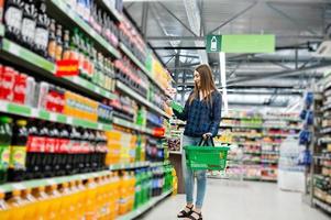 femme commerçante regardant les étagères du supermarché. portrait d'une jeune fille dans un magasin de marché tenant un panier vert et une bouteille de boisson gazeuse. photo