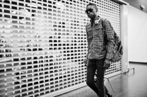 homme afro-américain en chemise à carreaux, lunettes de soleil et jeans avec valise et sac à dos. voyageur noir en duty free. photo