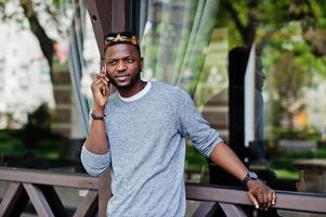 élégant garçon afro-américain sur un pull gris et des lunettes de soleil noires posées dans la rue et parlant au téléphone. mec noir à la mode. photo