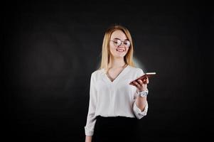 portrait en studio d'une femme d'affaires blonde à lunettes, chemisier blanc et jupe noire regardant un téléphone portable sur fond sombre. femme réussie et concept de fille élégante. photo