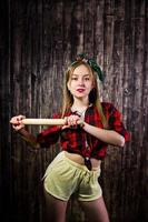 jeune femme au foyer drôle en chemise à carreaux et short jaune style pin up avec rouleau à pâtisserie de cuisine sur fond en bois. photo