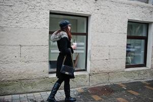 fille mexicaine bouclée en casquette en cuir et tasse de café en plastique à portée de main marchant dans les rues de la ville. photo