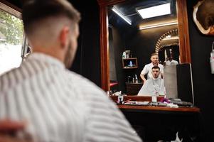 jeune homme barbu se coupe les cheveux par un coiffeur assis sur une chaise au salon de coiffure. âme de barbier. photo