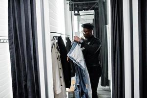 élégant homme afro-américain décontracté à la veste en jean et au béret noir au magasin de vêtements de cabine d'essayage changer de pardessus. photo