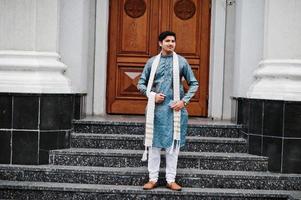 l'homme indien porte des vêtements traditionnels avec un foulard blanc posé à l'extérieur contre la porte du bâtiment avec un téléphone portable à portée de main. photo