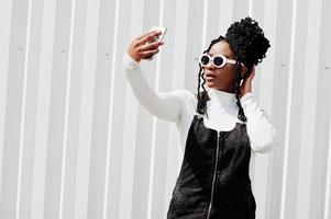 femme africaine en jupe salopette en jean, lunettes de soleil blanches posées contre un mur en acier blanc et faisant du selfie par téléphone. photo