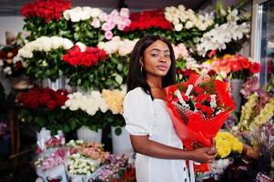 belle fille afro-américaine en robe blanche tendre avec bouquet de fleurs dans les mains debout sur fond floral dans le magasin de fleurs.fleuriste femme noire. photo