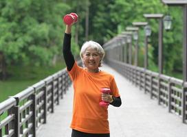 une femme asiatique à la retraite portant un t-shirt orange fait de l'exercice à l'extérieur, court, soulève des haltères, s'étire. photo