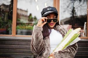 femme afro-américaine à la mode en casquette noire, lunettes de soleil et fourrure avec deux petits pains de baguette à portée de main. photo