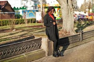 portrait d'une femme africaine aux cheveux bouclés portant un manteau noir à la mode et un col roulé rouge posant en plein air par une journée ensoleillée. photo