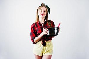 jeune femme au foyer drôle en chemise à carreaux et short jaune pin up style avec casserole et cuillère de cuisine isolé sur fond blanc. photo