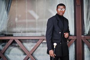 élégant gentleman afro-américain dans une élégante veste noire, tenant un bâton de marche rétro comme flacon de canne ou canne basculante avec poignée en boule de diamant doré. riche homme afro à la mode. photo