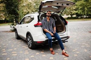 un homme arabe réussi porte une chemise rayée et des lunettes de soleil assis dans le coffre de sa voiture suv blanche avec téléphone portable. photo