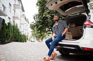 un homme arabe réussi porte une chemise rayée et des lunettes de soleil assis dans le coffre de sa voiture suv blanche. hommes arabes élégants dans les transports. photo