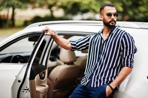 un homme arabe réussi porte une chemise rayée et des lunettes de soleil pose près de sa voiture suv blanche. hommes arabes élégants dans les transports. photo