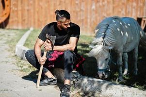 bûcheron homme barbe hipster arabe tenir la hache contre cheval poney. photo