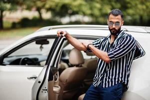 un homme arabe réussi porte une chemise rayée et des lunettes de soleil pose près de sa voiture suv blanche. hommes arabes élégants dans les transports. photo