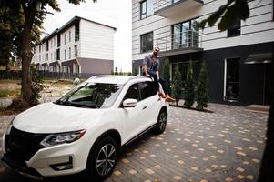 un homme arabe réussi porte une chemise rayée et des lunettes de soleil assis sur le toit de sa voiture suv blanche. photo