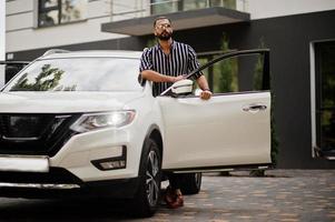 un homme arabe réussi porte une chemise rayée et des lunettes de soleil pose près de sa voiture suv blanche. hommes arabes élégants dans les transports. photo