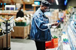 élégant homme afro-américain décontracté à la veste en jean et au béret noir tenant le panier et le lait à portée de main, debout près du réfrigérateur et faisant ses courses au supermarché.
