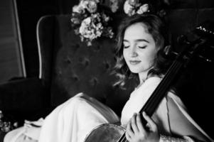 jolie jeune musicienne gilrl en robe blanche avec contrebasse assise sur un canapé marron vintage. photo